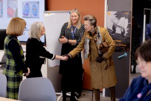 A woman in a brown coat shakes hands with another woman dressed in black, two other women look on